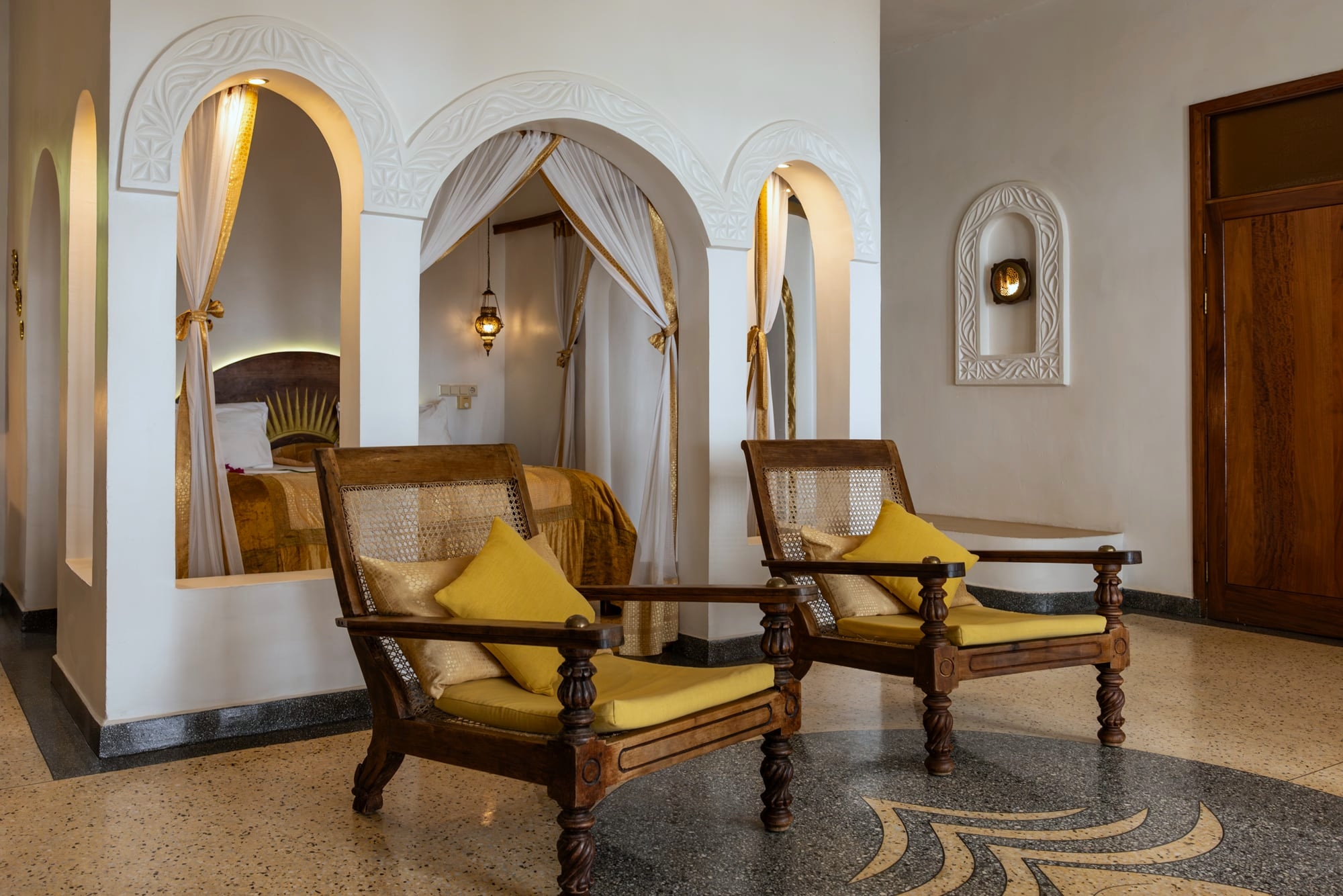 Jua: a draped bed with carved chairs and a writing table against white walls.