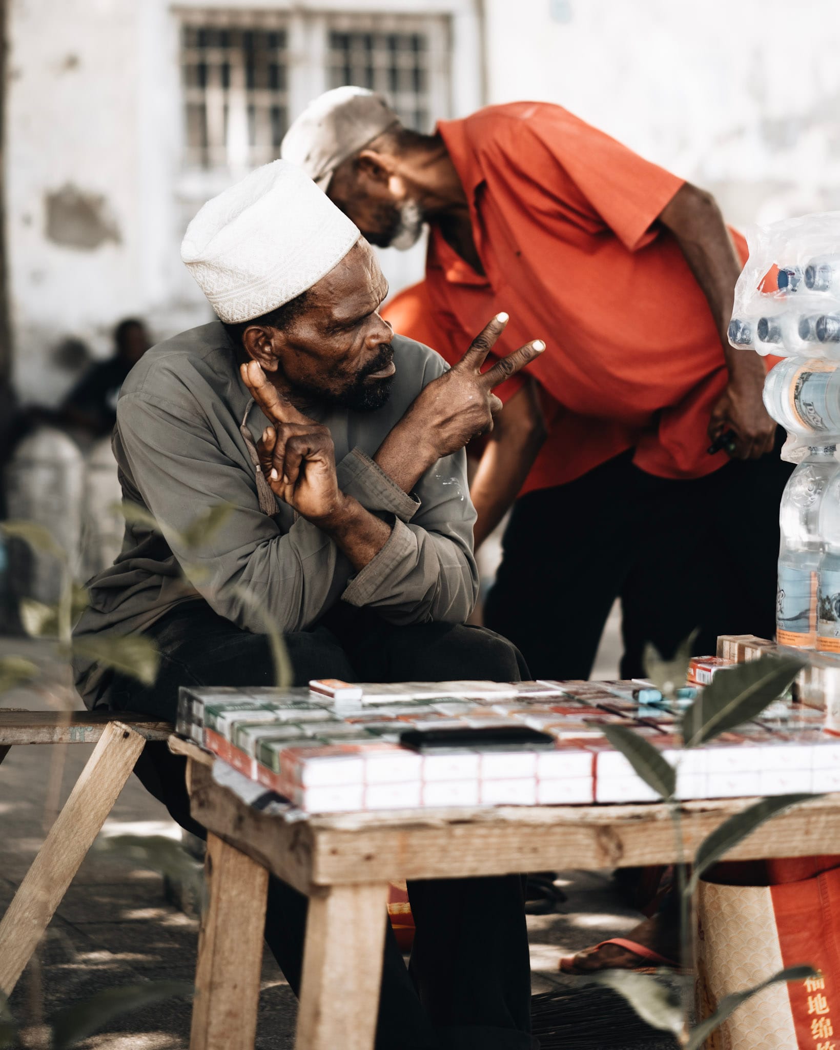 Two men in traditional attire at a Zanzibar market stall, one gesturing with his hands